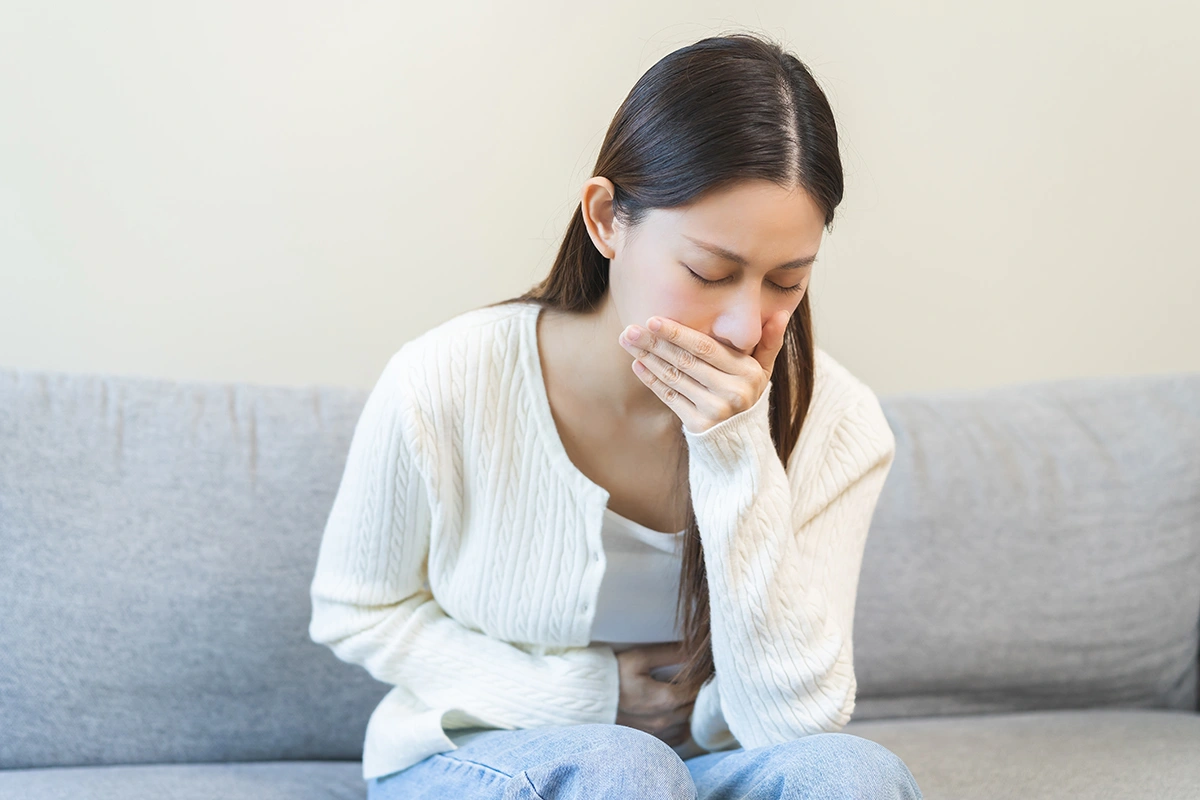 Woman sitting on a sofa holding her stomach and covering her mouth while experiencing nausea. 