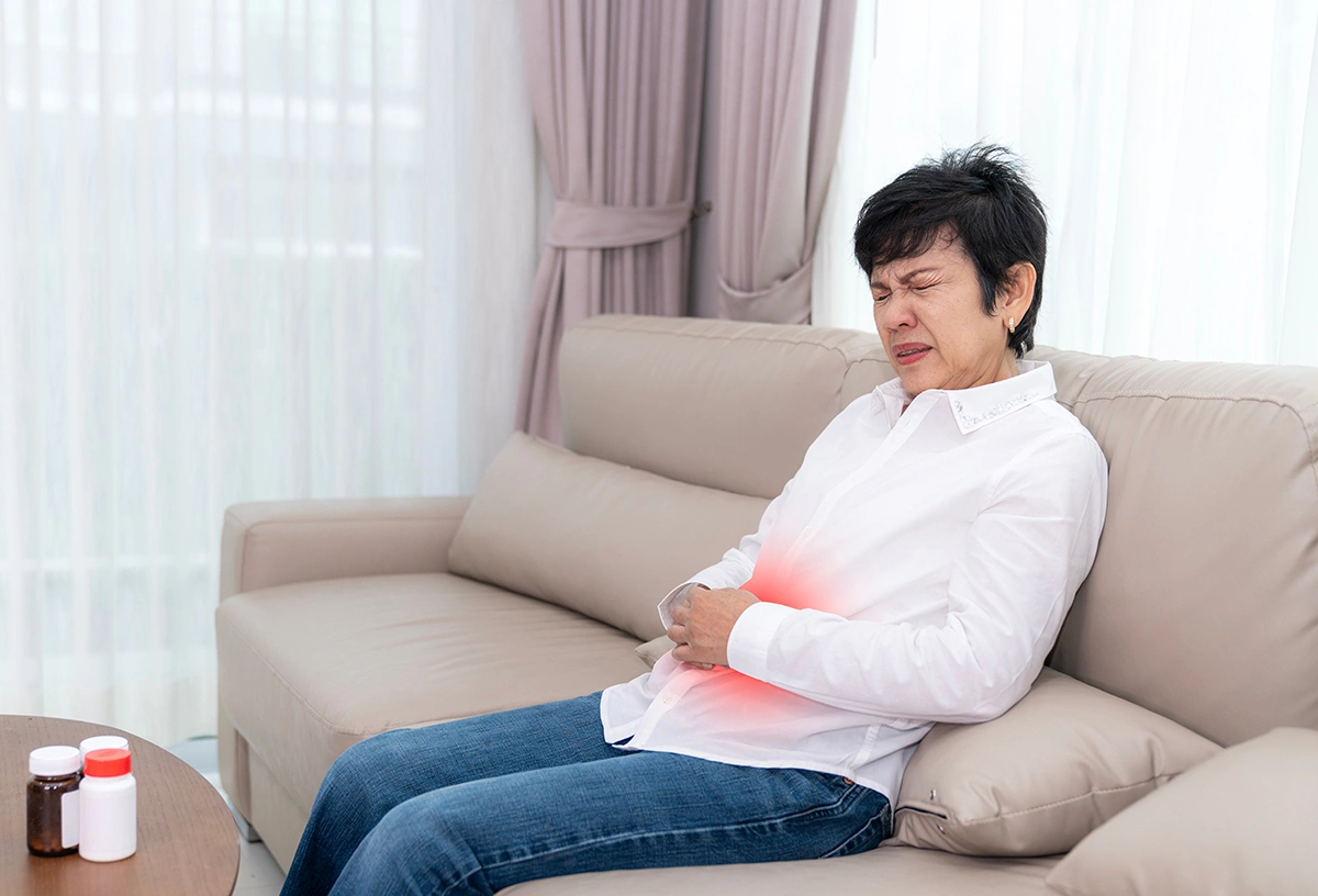 Woman sitting on a sofa leaning backwards and clutching her stomach while experiencing abdominal pain. 